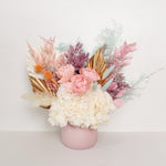 A mix of preserved flowers and silk flowers of hydrangea, Italian ruscus, billy button, misty, peony, bunny tail and palm in a in pink vase.